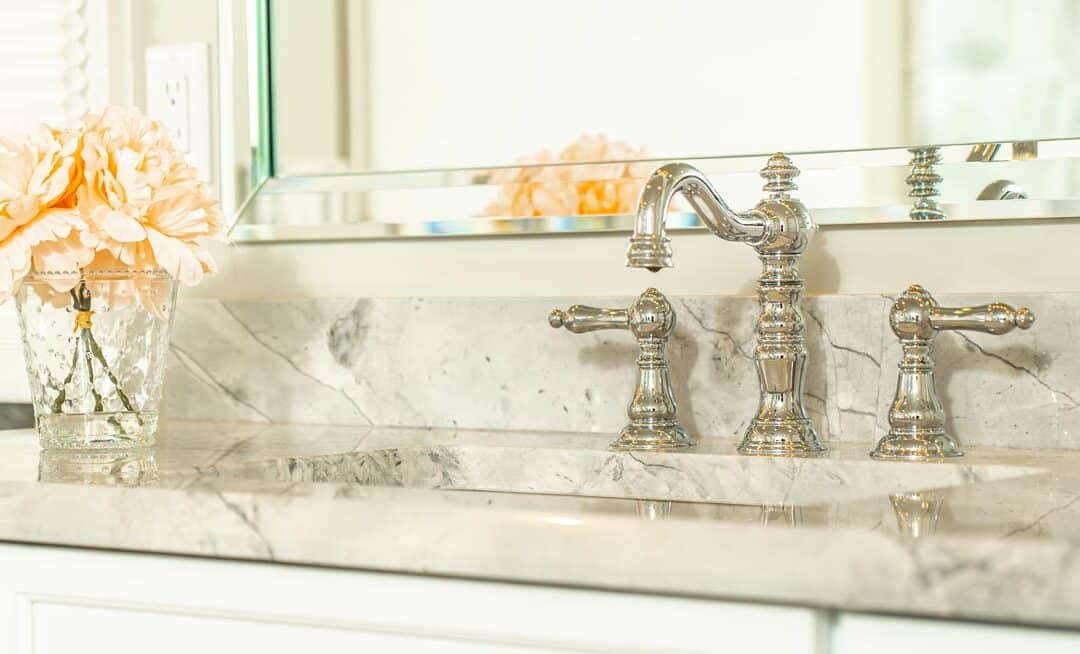 Classic chrome faucet on a white marble bathroom vanity next to a vase of peach flowers.