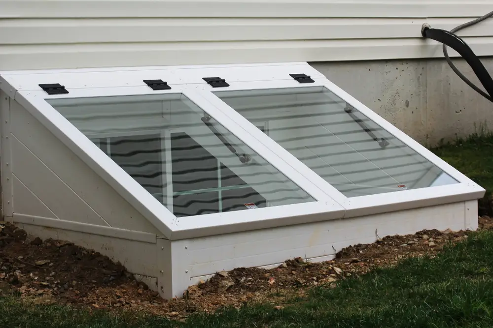 White sloped basement egress window well cover with glass panels outside a house.
