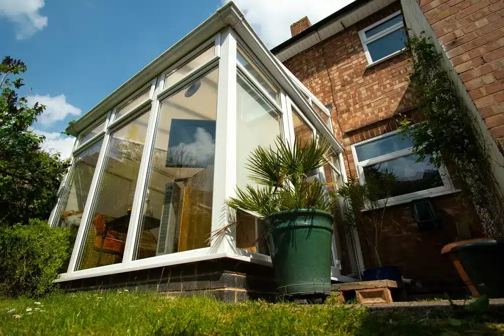 Exterior low-angle view of a modern glass-walled sunroom addition attached to a two-story brick main house.