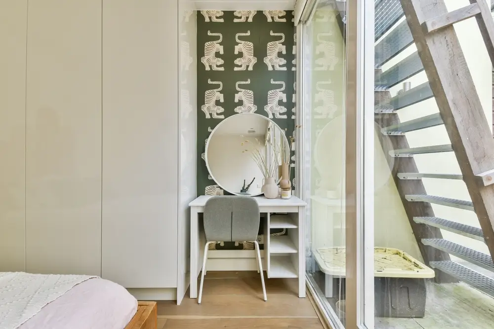 Compact bedroom in-law suite nook featuring built-in wardrobes, a small vanity desk with a round mirror, and floor-to-ceiling glass doors.