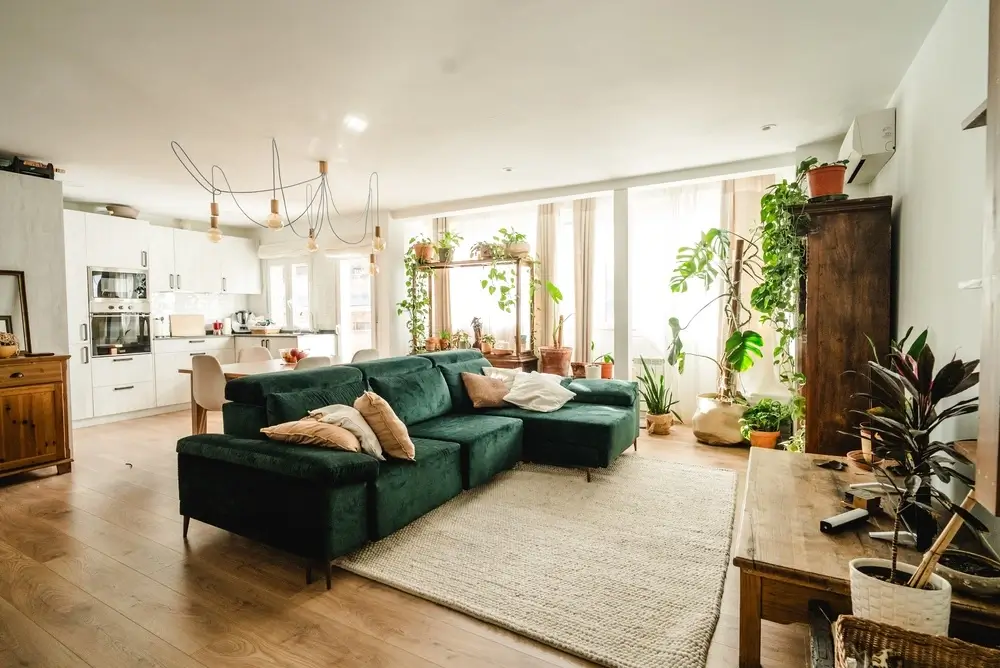 Open-concept in-law suite living room featuring a green sectional sofa, wood flooring, a full white kitchen, and a wall-mounted climate control unit.