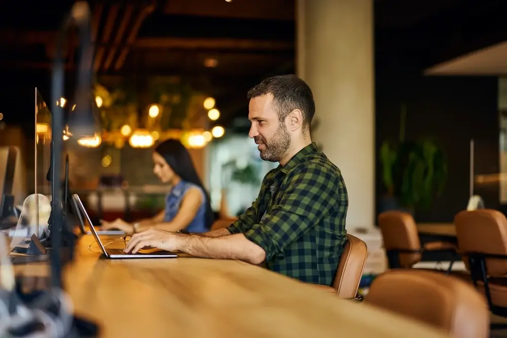 Man working on a laptop at a shared wooden table in a co-working space.